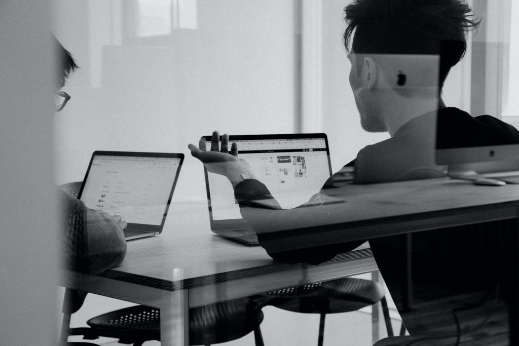 Mustavalkoinen kuva, jossa kaksi henkilöä istuu pöydän äärellä ja katsoo tietokoneita.